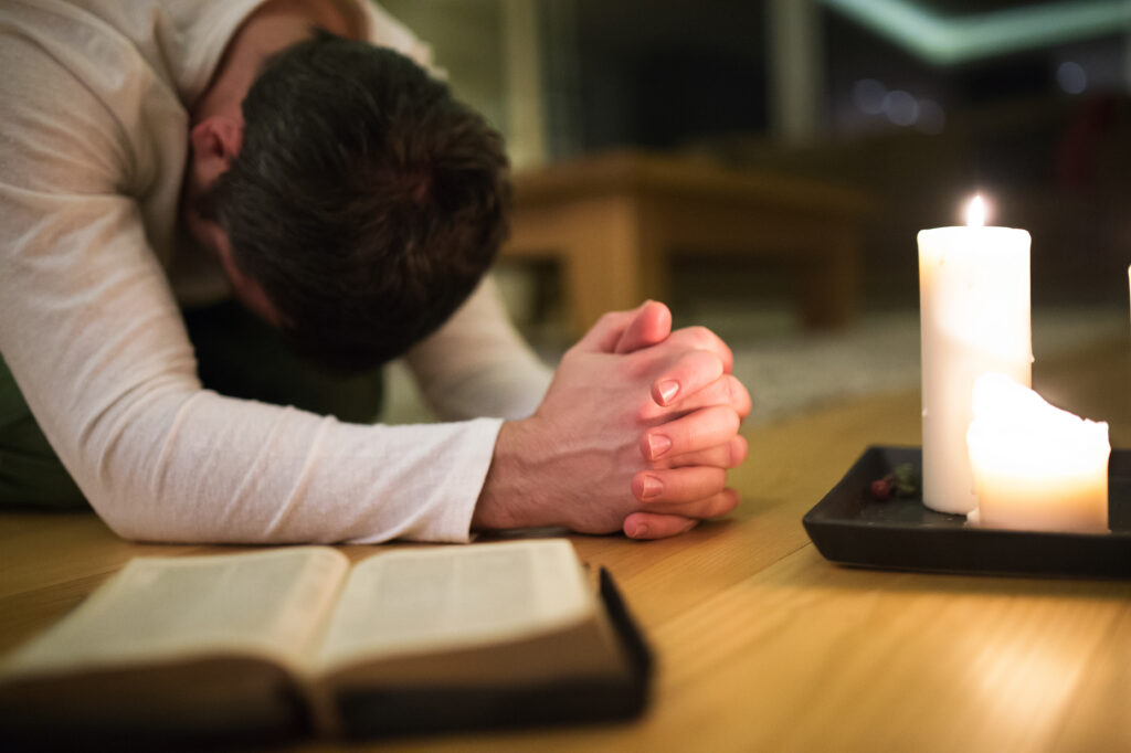 graphicstock unrecognizable young man praying kneeling on the floor hands clasped together bible and burning candles next to him close up rdM7VTOIMW