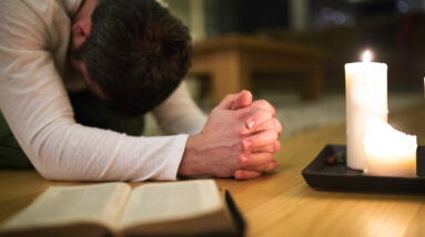 graphicstock unrecognizable young man praying kneeling on the floor hands clasped together bible and burning candles next to him close up rdM7VTOIMW